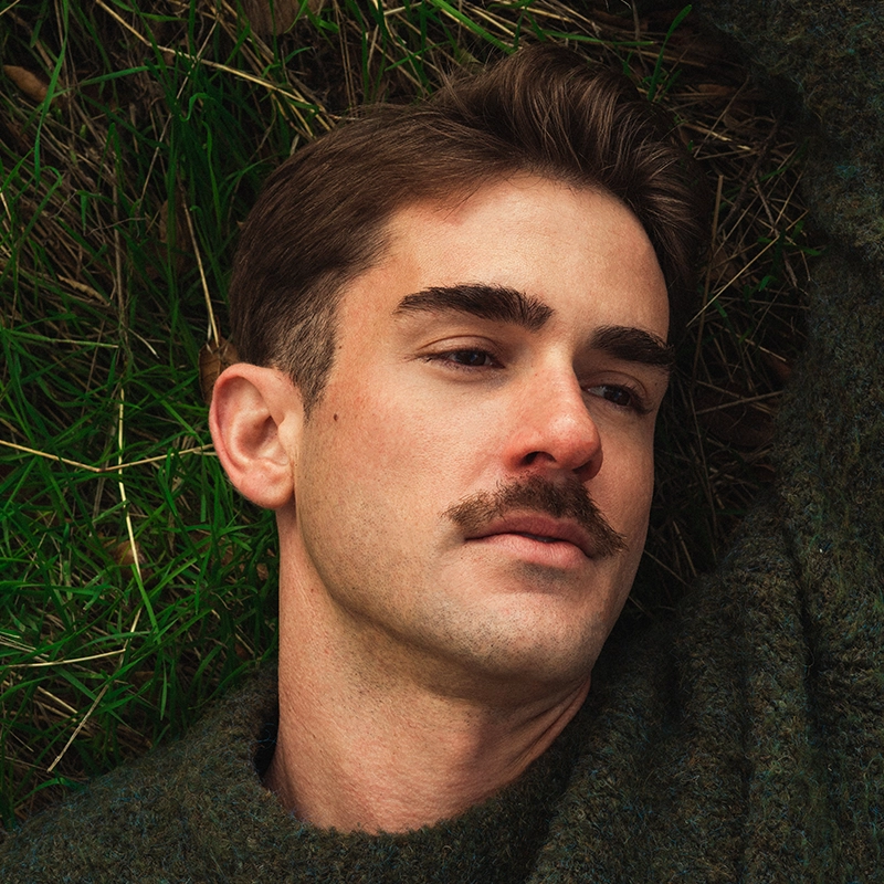 A young man with short brown hair, a thick mustache, and thick eyebrows lies on green grass. Wearing a dark, textured sweater, he gazes thoughtfully to the side amid brown leaves—captured in the serene style of Jana Djenne Jackson.