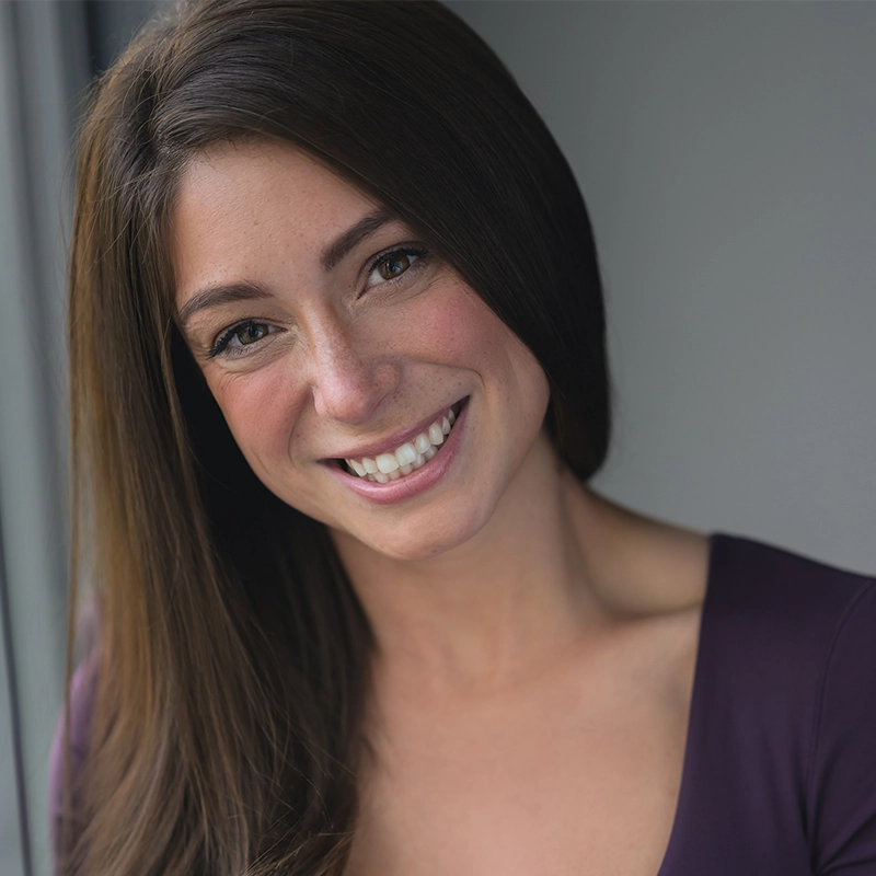 A woman with long brown hair smiles warmly, showing her teeth. Carissa Navarra wears a deep purple top and stands near a window with natural light on her face. The background is softly blurred gray.