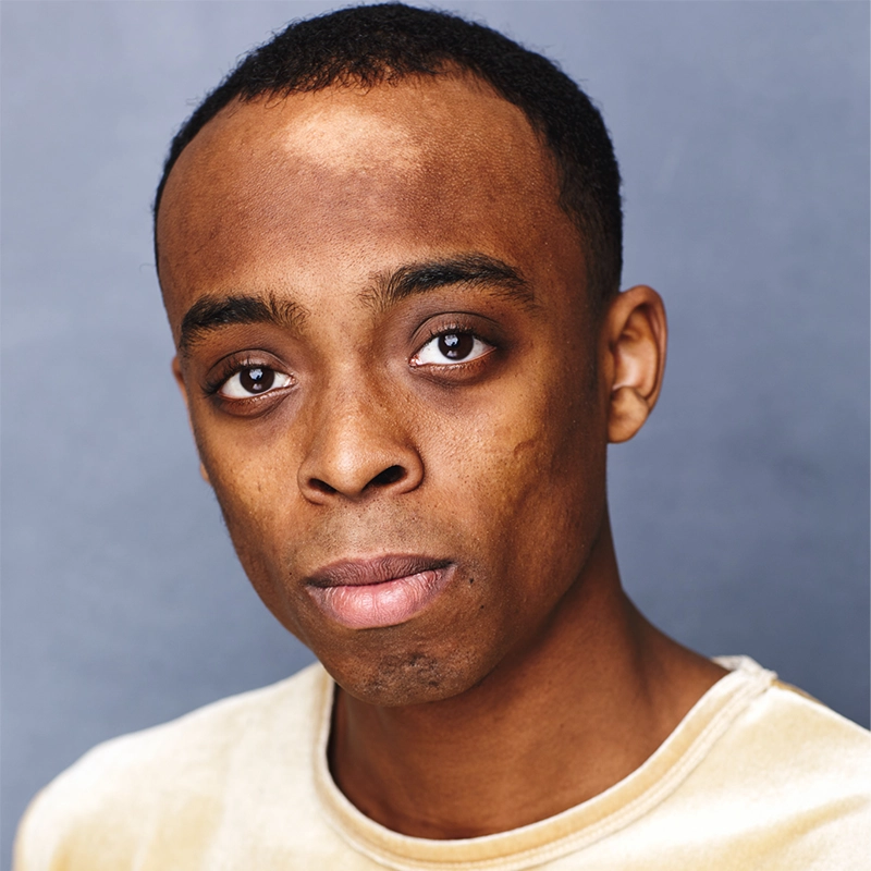 A young man with short black hair and brown eyes looks directly at the camera with a neutral expression. Styled by Ella Sodam Yoon, he wears a light beige shirt, posed against a plain, grayish-blue background under soft lighting.