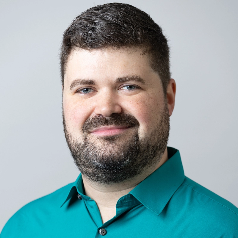 A man with short brown hair, a full beard, and blue eyes smiles slightly at the camera. He is wearing a teal button-up shirt and is posed against a plain, light gray background.