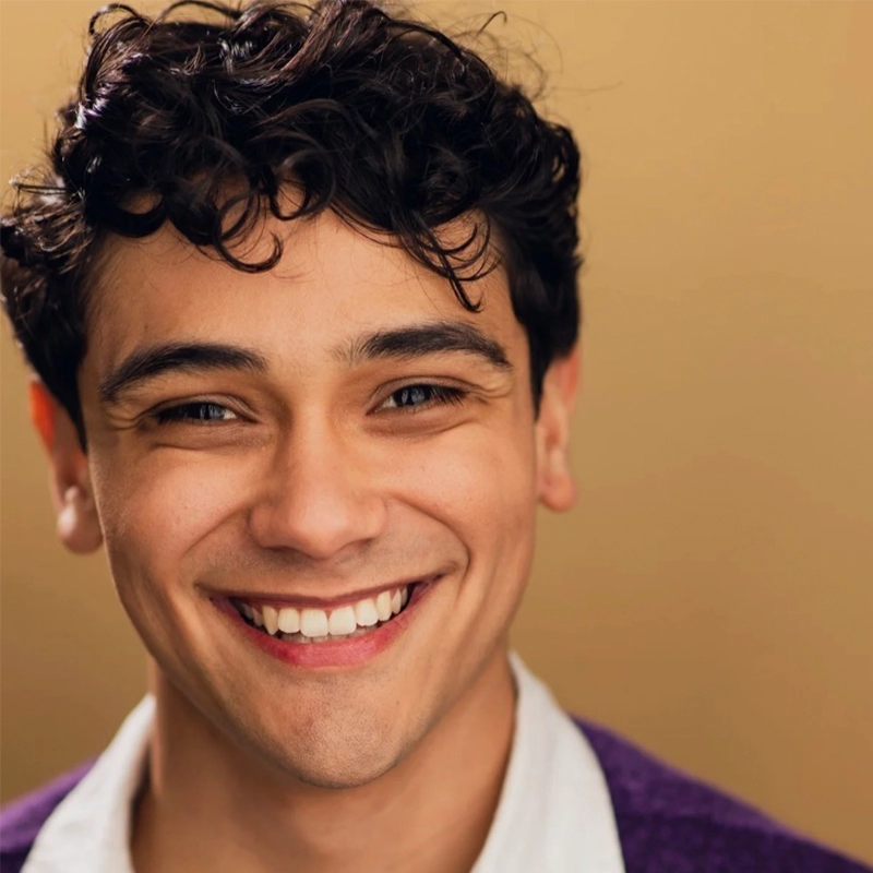 A young man with curly dark hair smiles warmly at the camera. He has light skin, brown eyes, and dimples. He wears a white collared shirt under a purple sweater, reminiscent of Katie Lemmen’s classic and approachable style. The background is a soft, solid beige color.