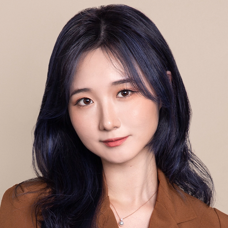 Ella Sodam Yoon, a young woman with long, dark wavy hair and fair skin, wears a brown blazer and a delicate pearl necklace. She faces the camera, smiling gently against a neutral beige background.