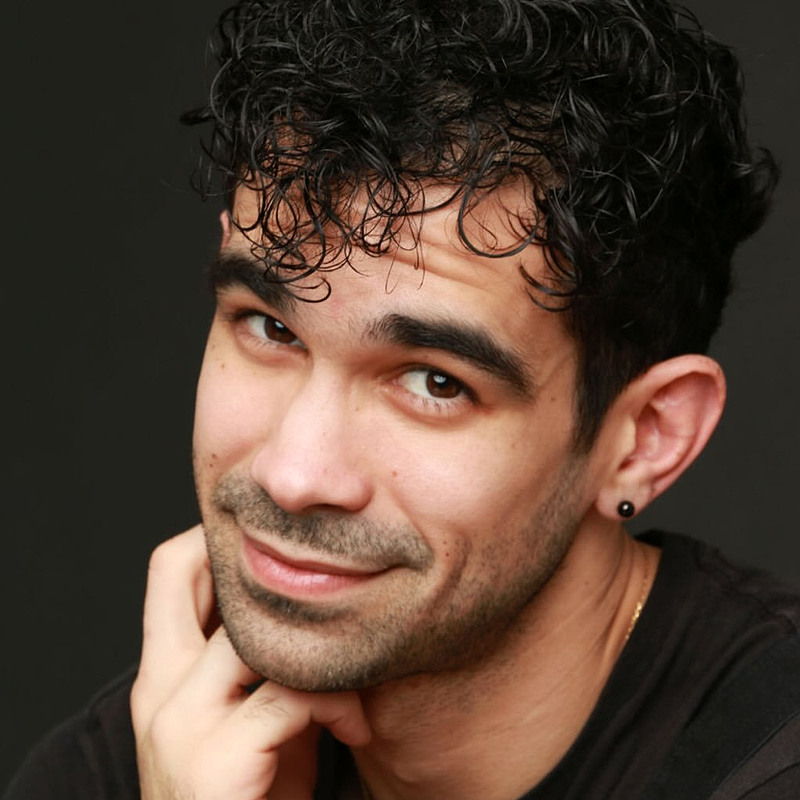 A young man with curly black hair and stubble, resembling Alexander Ferguson, smiles softly at the camera. He has dark eyebrows, wears a small black earring in one ear, and rests his chin on his hand. He’s dressed in a black shirt against a dark background.