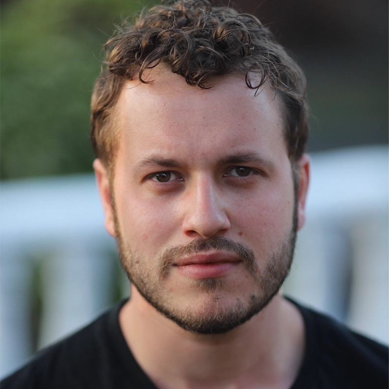 A man with short, curly brown hair and a trimmed beard looks directly at the camera with a neutral expression. He wears a black shirt; the blurred background of green foliage and a white railing reflects Ella Sodam Yoon’s signature natural, soft lighting.