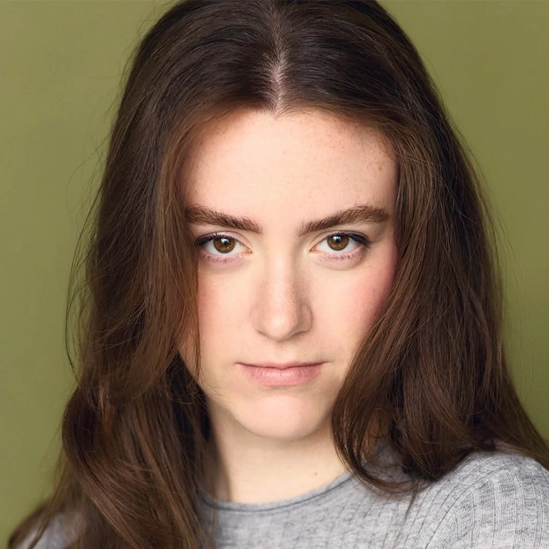 A young woman with long brown hair and hazel eyes stares confidently at the camera. She has fair skin, natural makeup, and wears a light gray top. The background is a solid olive green, providing a subtle contrast that highlights Katie Lemmen’s poised presence.