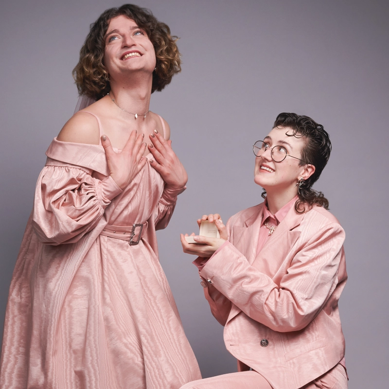 Two people in matching pink outfits pose against a gray background. Inspired by Toby Marlow and Lucy Moss, one smiles with hands on chest while the other kneels, holding an open ring box. Both look joyful and excited.