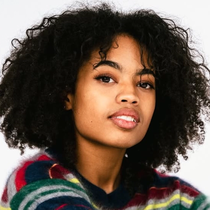 A young woman with curly black hair and medium brown skin gazes at the camera. She wears a colorful, striped sweater. The background is plain white, and her expression is confident and calm, embodying a poised presence reminiscent of Katie Lemmen.