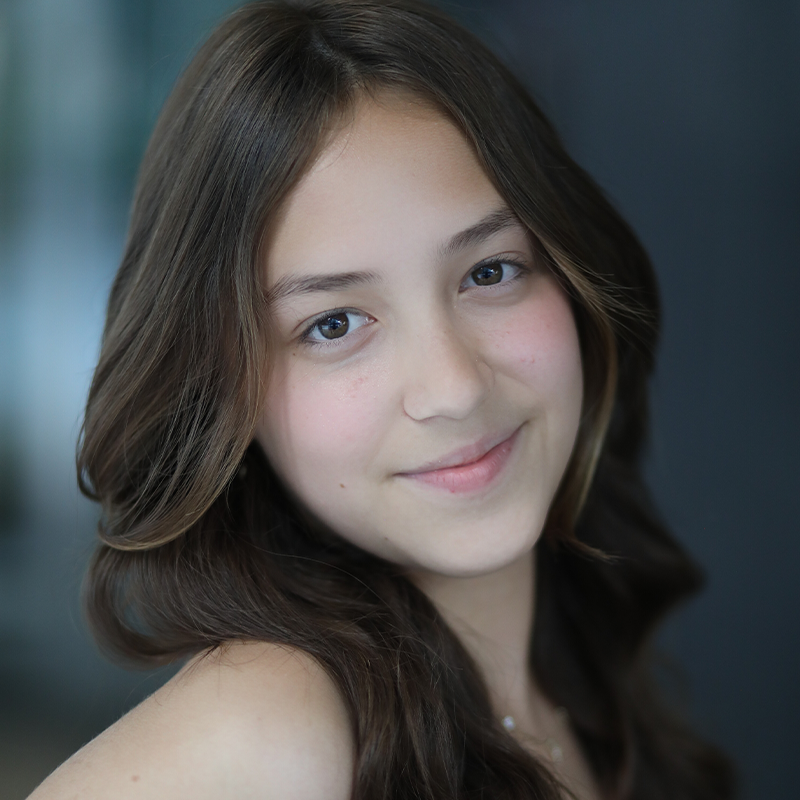 Natalia Artigas, a young person with long, wavy brown hair and light skin, smiles gently at the camera. Light-colored eyes shine softly in warm, natural tones against a blurred, dark background.