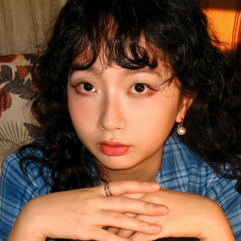 Ella Sodam Yoon, a young woman with long curly black hair and pearl earrings, looks directly at the camera. She wears a blue plaid shirt and rests her chin on her folded hands. Warm light and a floral-patterned couch are visible behind her.