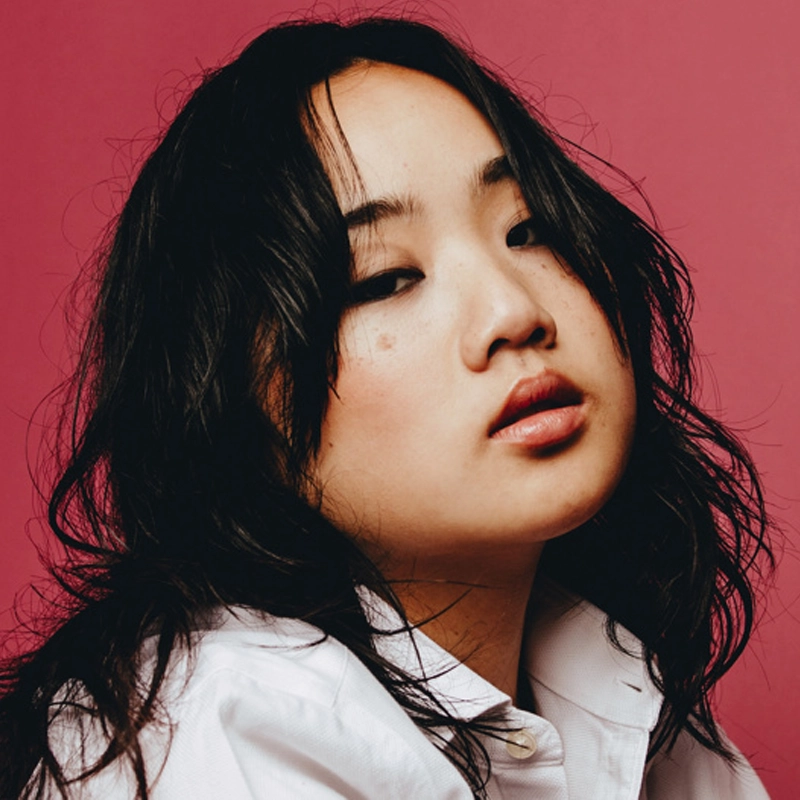 Helen J Shen, a woman with long, wavy black hair, poses against a pink background. She has light skin, natural makeup, and full lips. Wearing a white collared shirt, she gazes confidently at the camera with a slightly tilted head.