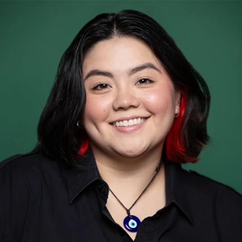 A person with medium-length dark hair and red tips smiles at the camera. They wear a black shirt and a necklace with a blue eye pendant. The background is a solid dark green. Their cheeks are slightly rosy, and they have dimples.