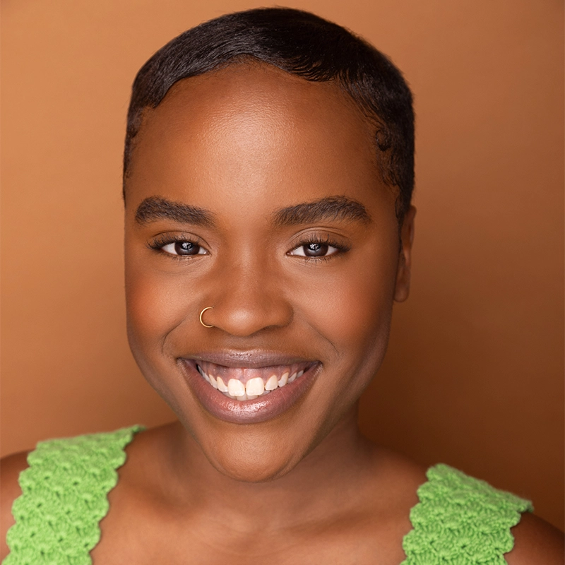 A smiling person with smooth dark skin and short, neatly styled hair poses against a warm brown background. They wear a green crochet top and a small gold nose ring on the left nostril, showcasing bright, even teeth and natural makeup.