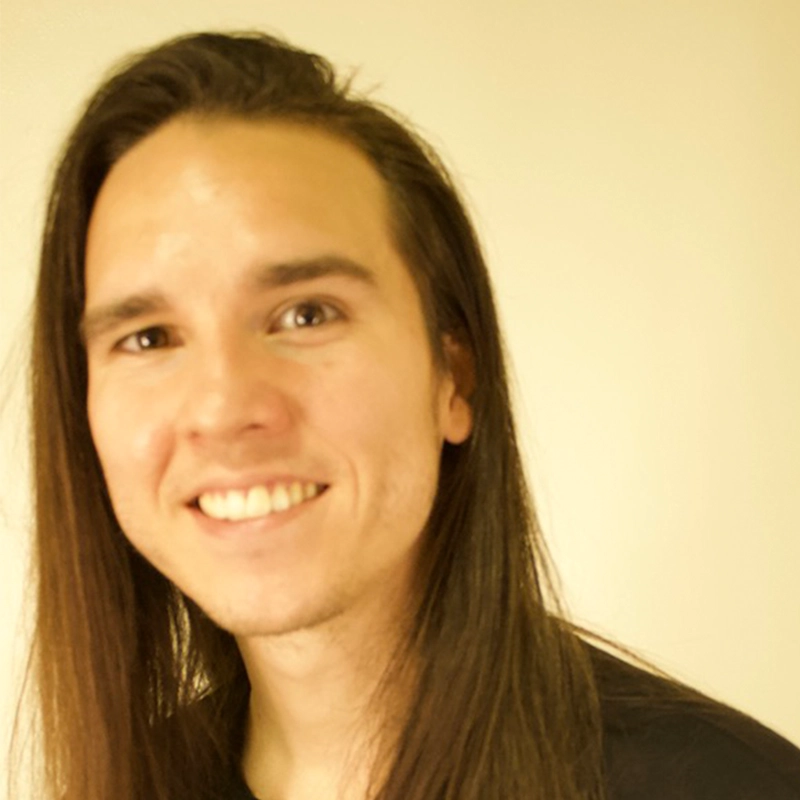 Ella Sodam Yoon, with long straight brown hair and light skin, smiles at the camera. She wears a black shirt and is posed in front of a plain, light-colored background. The lighting is soft, lending the image a warm tone.
