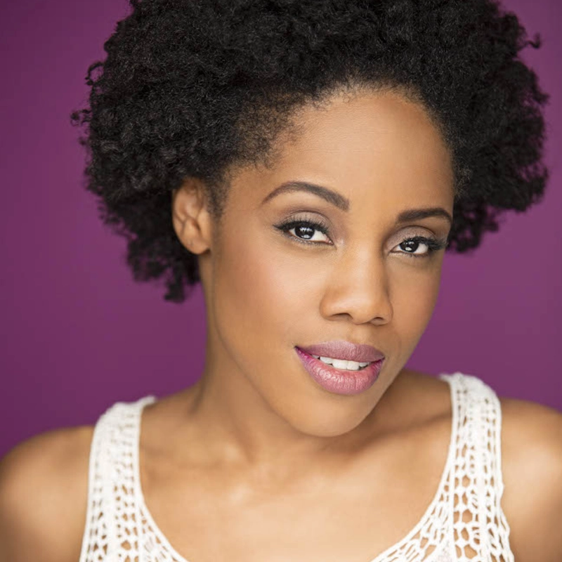 Zuri Washington, with natural, curly black hair, smiles softly at the camera. She wears light makeup and a white, sleeveless crochet top. The vibrant purple background highlights her smooth, warm brown skin in perfect lighting.