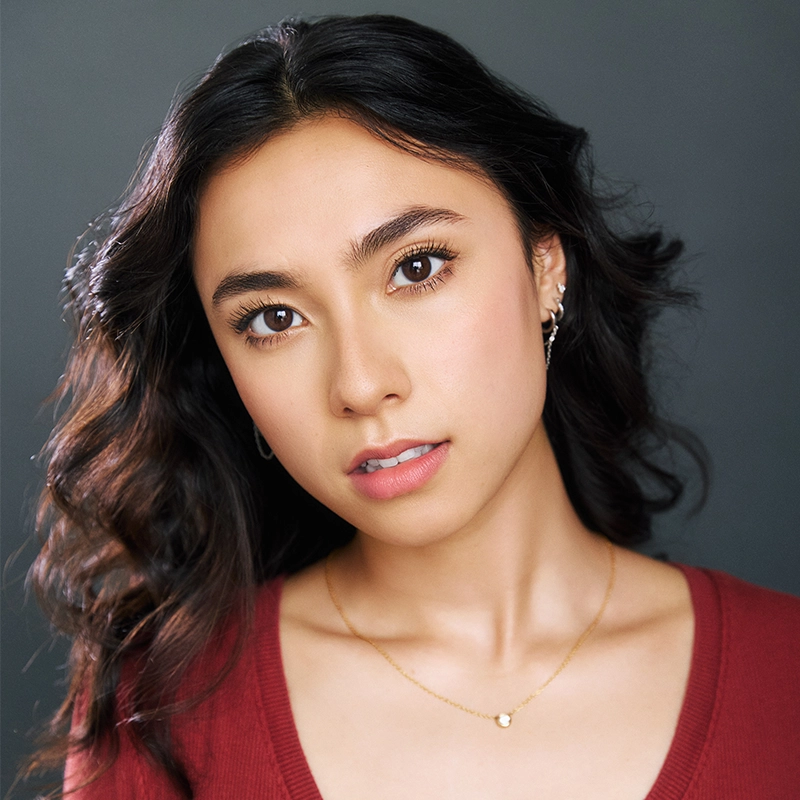 A young woman with wavy dark hair and brown eyes gazes calmly at the camera. She wears natural makeup, a red top, small hoop earrings, and a gold necklace with a small pendant. The background is plain and gray, softly lit.