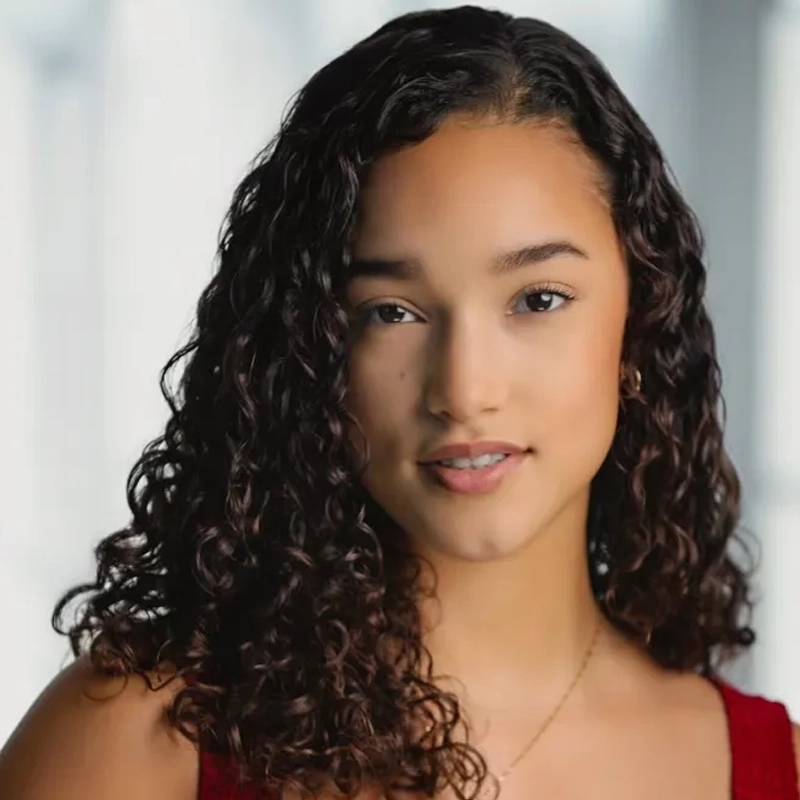Headshot of Isa Antonetti, a young woman with long, dark curly hair looking toward the camera. She has a neutral expression and is wearing a red top.