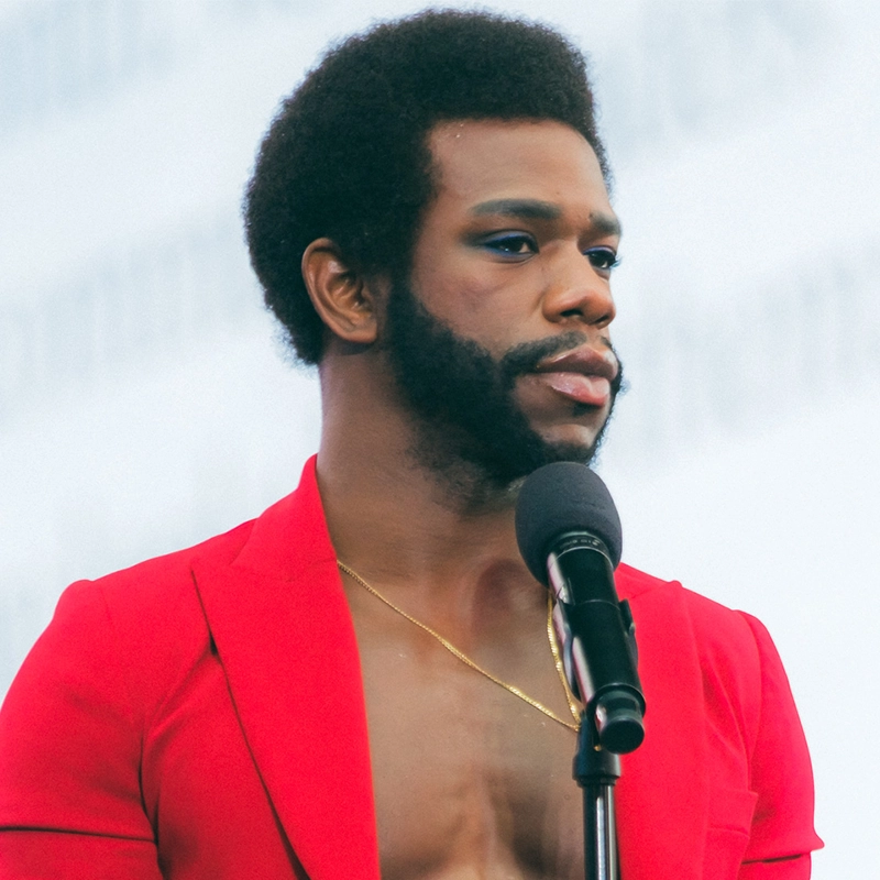A person with short, curly hair, a full beard, and sideburns stands at a microphone. They wear a bright red open jacket with no shirt, showing a gold necklace and bare chest. The confident pose and bold style are reminiscent of Antwayn Hopper. The background is light and out of focus.