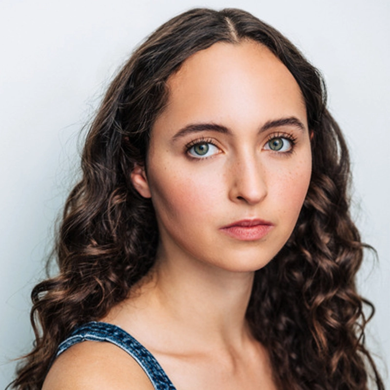 Young woman with long, dark, curly hair and green eyes looks directly at the camera with a neutral expression. She wears a blue tank top and is posed against a plain, light background. Her skin is fair with subtle makeup and faint freckles, reminiscent of the natural aesthetic often captured in Joey LaVarco’s portraits.