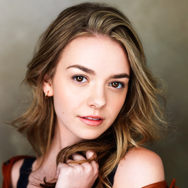 A young woman with wavy blonde hair, brown eyes, and light skin looks into the camera. She wears a dark top and a small earring, gently holding her hair and jacket. The background is softly blurred in neutral tones, capturing an aesthetic reminiscent of a Joey LaVarco portrait.