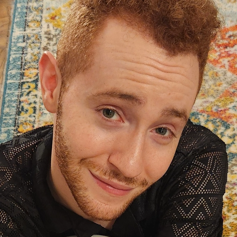 A young person with curly red hair and a short beard smiles at the camera. Matthew Berzon wears a black patterned shirt and sits on a colorful, patterned rug with shades of blue, yellow, and orange in the background.