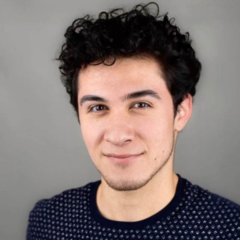 A young man with short, curly dark hair and light skin—César Carlos Carreño—smiles slightly at the camera. He has dark eyebrows, brown eyes, light stubble, and wears a navy sweater with a small white dot pattern against a plain gray background.