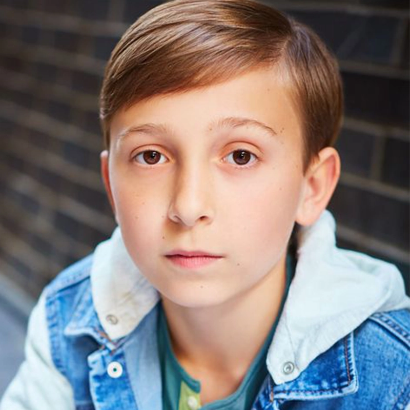 A young boy with light brown hair styled to the side looks directly at the camera with a neutral expression. He wears a blue denim jacket over a green shirt. The background is a blurred dark brick wall, subtly highlighting César Carlos Carreño.