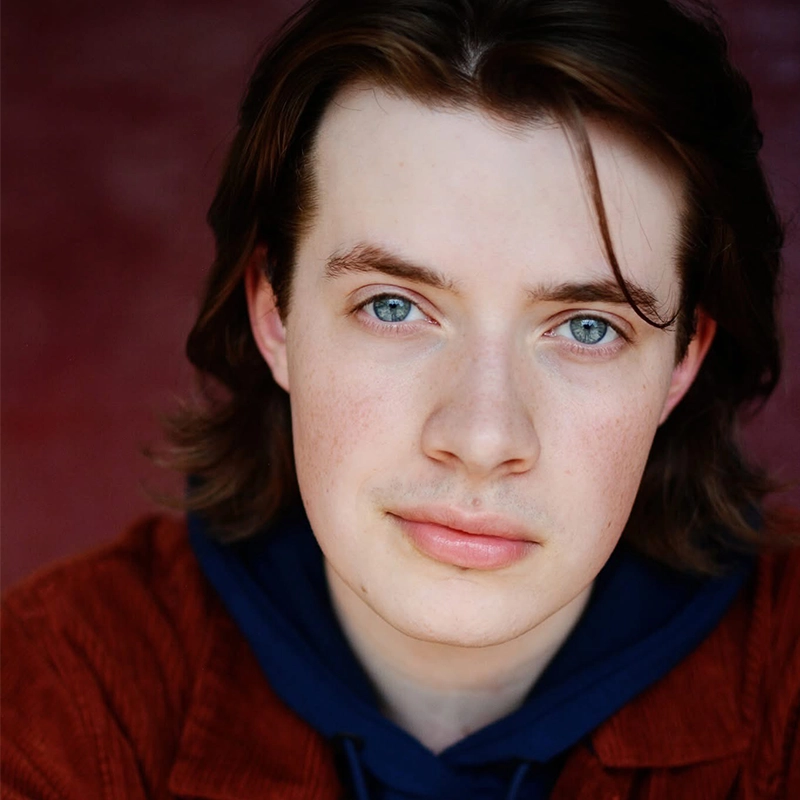 A young person with fair skin, blue eyes, and medium-length brown hair looks directly at the camera. They wear a rust-colored jacket over a navy hoodie, with a calm, neutral expression and a blurred, dark red background.