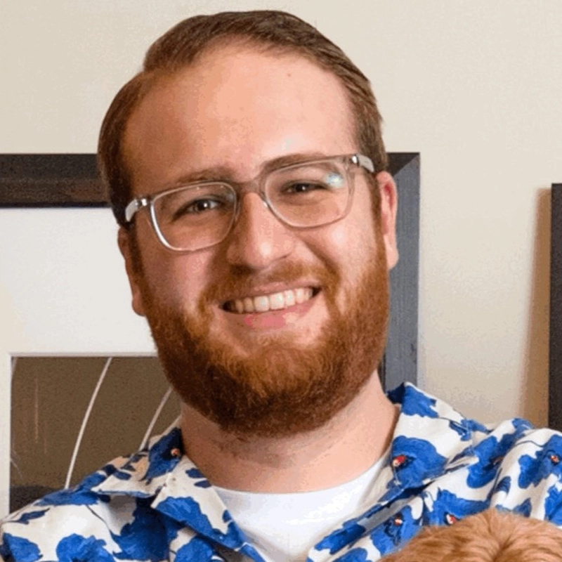 A smiling man, James Feinberg, with short brown hair, a trimmed beard, and glasses wears a white shirt with a blue floral pattern. He stands indoors in front of a beige wall with framed artwork partially visible behind him.