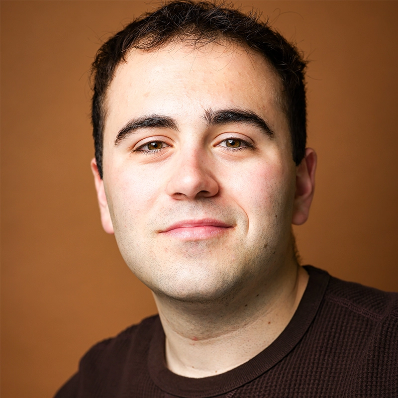 A man with short dark hair and fair skin smiles softly at the camera. He is wearing a dark brown textured long-sleeve shirt. The background is a plain, warm brown color, giving the portrait a friendly and approachable feel.