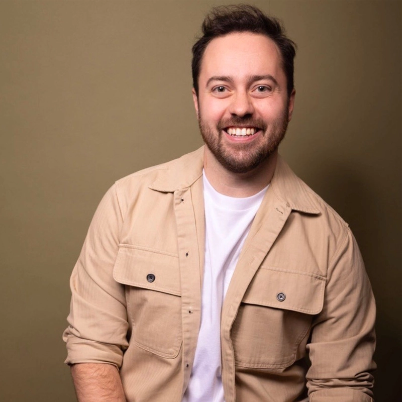 Matt Kurzyniec, a smiling man with short dark hair and a trimmed beard, wears a beige button-up shirt over a white t-shirt. He is posed slightly angled to the left, arm bent, against a plain olive-green background.