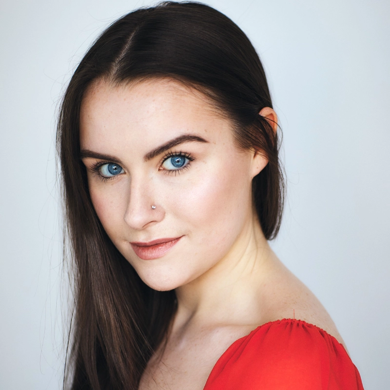 Grace Carey McGovern, a young woman with long straight brown hair and bright blue eyes, looks at the camera, smiling softly. She has a small nose piercing and wears a red off-the-shoulder top. The background is a plain, light color.