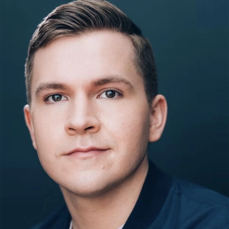 A young man with short, light brown hair styled neatly, fair skin, and blue eyes looks directly at the camera with a neutral expression. He wears a dark blue jacket. The background is a solid, dark teal color. This portrait features Jack B. Murphy.
