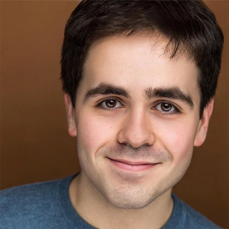 Noah Marlowe, a young man with short dark brown hair and fair skin, smiles gently. He has dark brown eyes, thick eyebrows, and wears a blue crew-neck shirt while posed against a plain brown background.