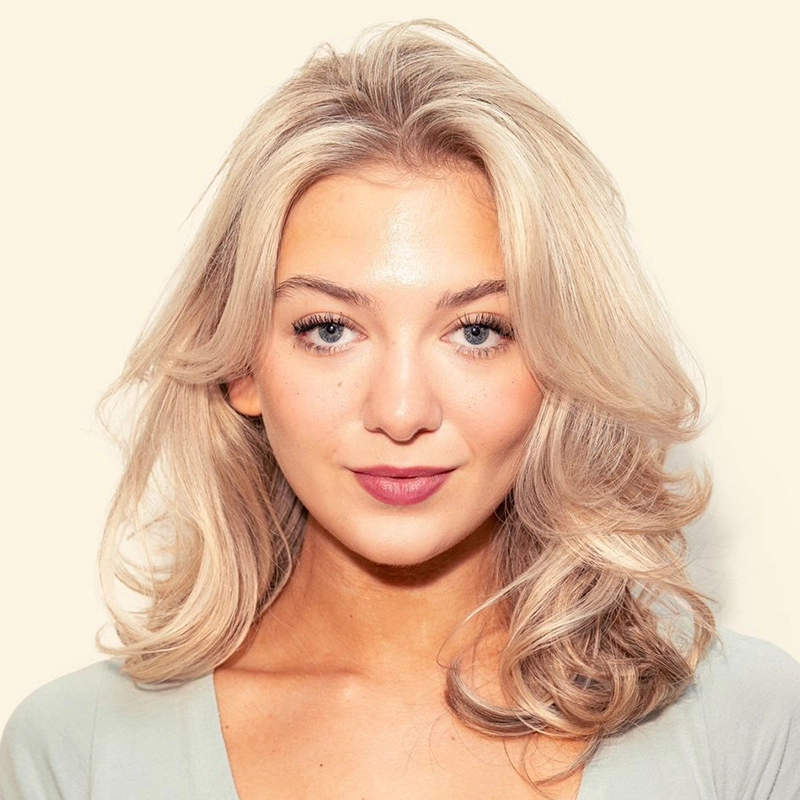 A young woman, Callie Henrysen, with wavy, shoulder-length blonde hair, blue eyes, and light skin stands against a cream background. She is wearing a pale top and has a calm, confident expression with neutral makeup and rose-colored lipstick.