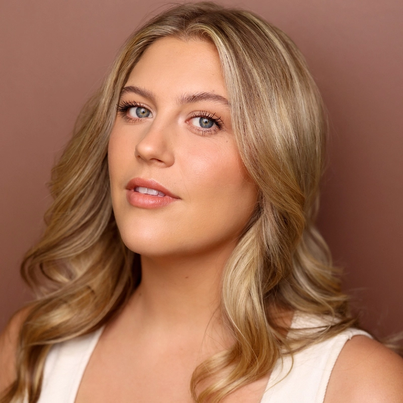 A woman with long, wavy blonde hair and fair skin poses against a muted pink background. She wears a white top, natural makeup, and soft pink lips. Her calm, confident expression reflects Bella Coppola's effortless elegance as she looks slightly off-camera.