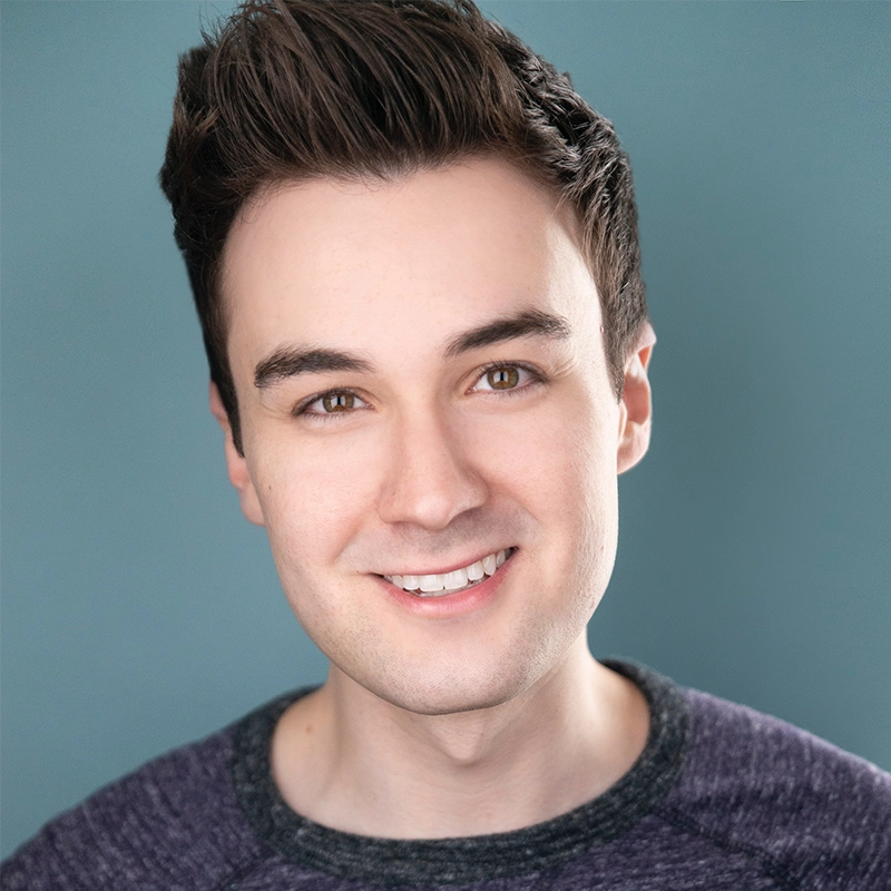 A young man with fair skin, short dark brown hair styled up, and brown eyes is smiling at the camera. He is wearing a dark, textured crew-neck sweater. The soft, even lighting and teal blue background evoke a portrait style seen in Natalia Vivino's works.