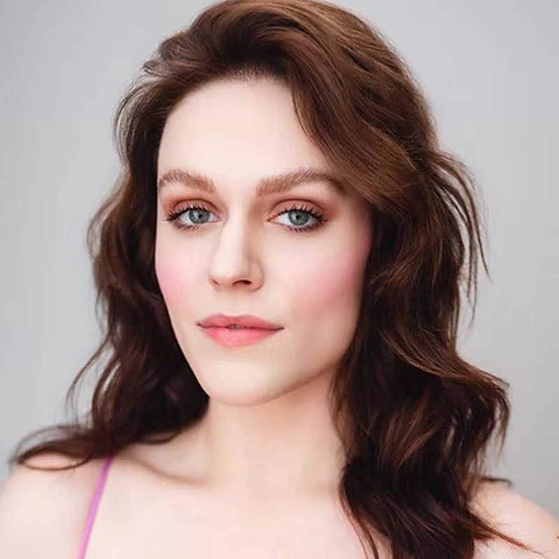 A woman with fair skin, long wavy brown hair, and blue eyes faces the camera against a plain light background. Wearing natural makeup and a pink strap top, Kylie Heyman exudes a calm and confident expression.