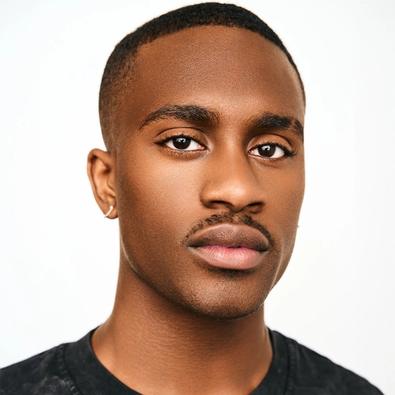 A young man named Jacobi with short hair and a trimmed mustache looks directly at the camera. He has brown skin, expressive dark eyes, and wears a small hoop earring in his left ear. He is dressed in a black shirt against a plain white background.