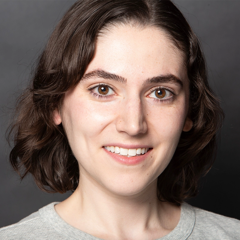 A young woman with short, wavy brown hair smiles at the camera. She has fair skin, brown eyes, and thick eyebrows. Natalia Vivino is wearing a light grey top and is posed against a plain dark grey background.