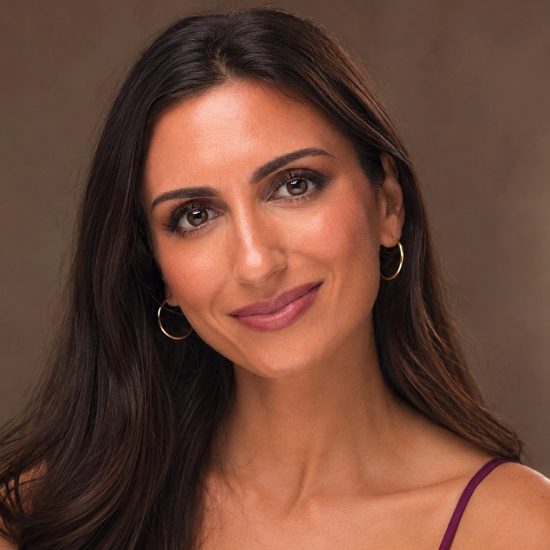 A woman with long dark hair and medium skin tone smiles gently at the camera. Natalia Vivino wears gold hoop earrings, subtle makeup, and a maroon camisole. The background is softly blurred in warm brown tones.