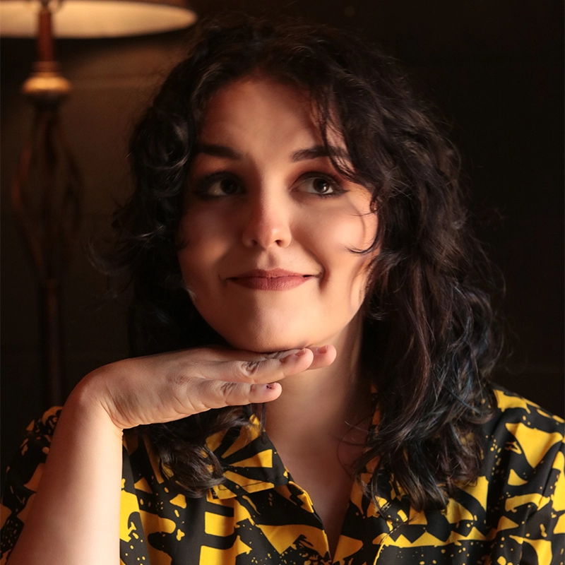 Analisa Reynoso, with wavy dark hair, smiles softly, resting her chin on her hand. She wears a yellow and black patterned shirt and sits in warmly lit surroundings, with a lamp and dark wall in the background.