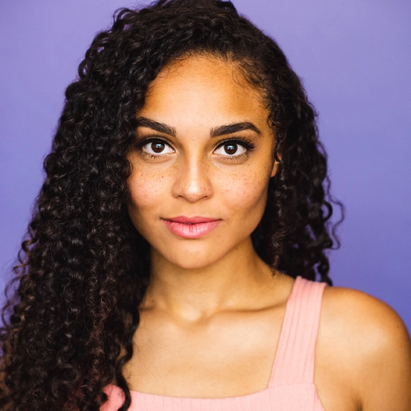 Jisel Soleil Ayon, a young woman with medium skin tone, curly dark hair, and brown eyes, gazes confidently at the camera. She wears a sleeveless light pink top and subtle makeup against a solid light purple background.