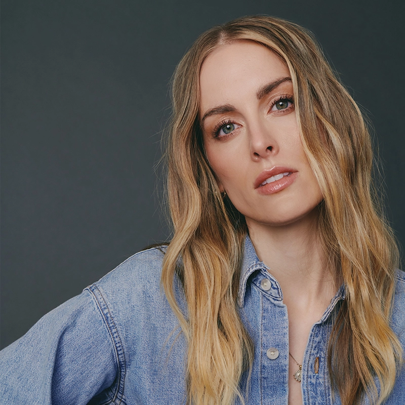 Bailey McCall, with long, wavy blonde hair and fair skin, wears a light blue denim shirt. She looks directly at the camera with a neutral expression against a plain, dark gray background.
