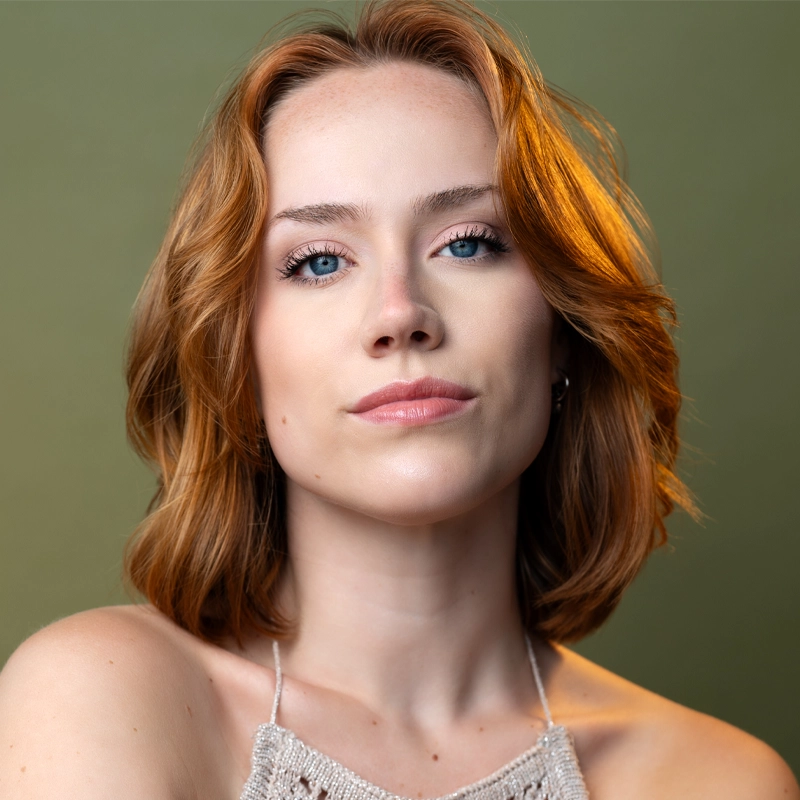 Maggie Bergman, a young woman with fair skin, blue eyes, and shoulder-length auburn hair, looks confidently at the camera. She wears natural makeup and a beige knit halter top, with warm light highlighting her hair against a softly blurred green background.