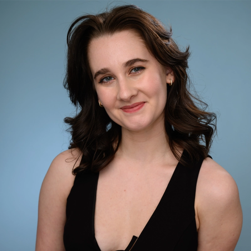 Emmarose Campbell, a young woman with fair skin and wavy dark brown hair, smiles gently at the camera. She wears a sleeveless black top and small gold hoop earrings, posed against a plain light blue background.