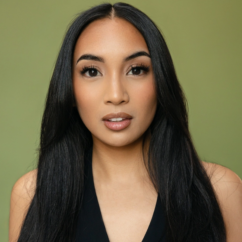 A woman with long, straight black hair and light brown skin looks directly at the camera. KC Dela Cruz wears natural makeup with defined brows and glossy lips, dressed in a sleeveless black top against a plain olive green background.