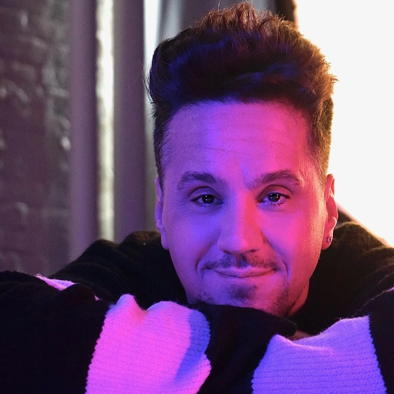 Henry Gainza, with styled dark hair and a trimmed beard, smiles gently, resting his chin on folded arms. He wears a black sweater with white stripes as pink and purple hues light up his face against a softly blurred background.