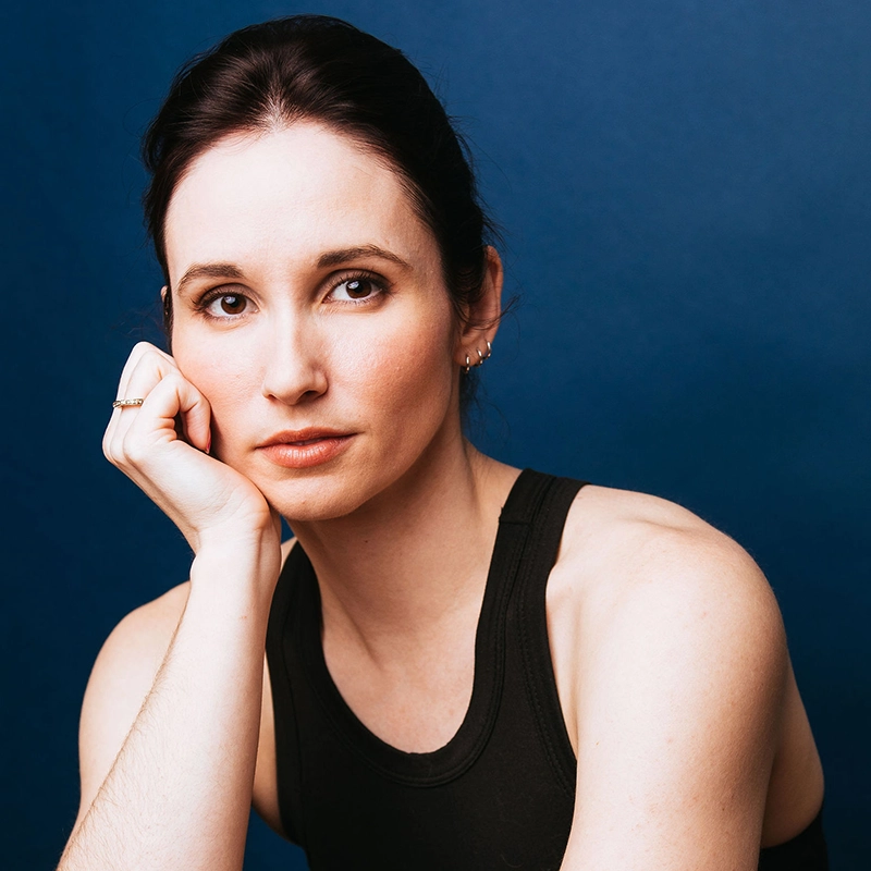 Bailey McCall, with fair skin and dark brown hair pulled back, rests her chin on her hand, gazing thoughtfully at the camera. She wears a black sleeveless top, a ring, and small earrings against a solid deep blue background.