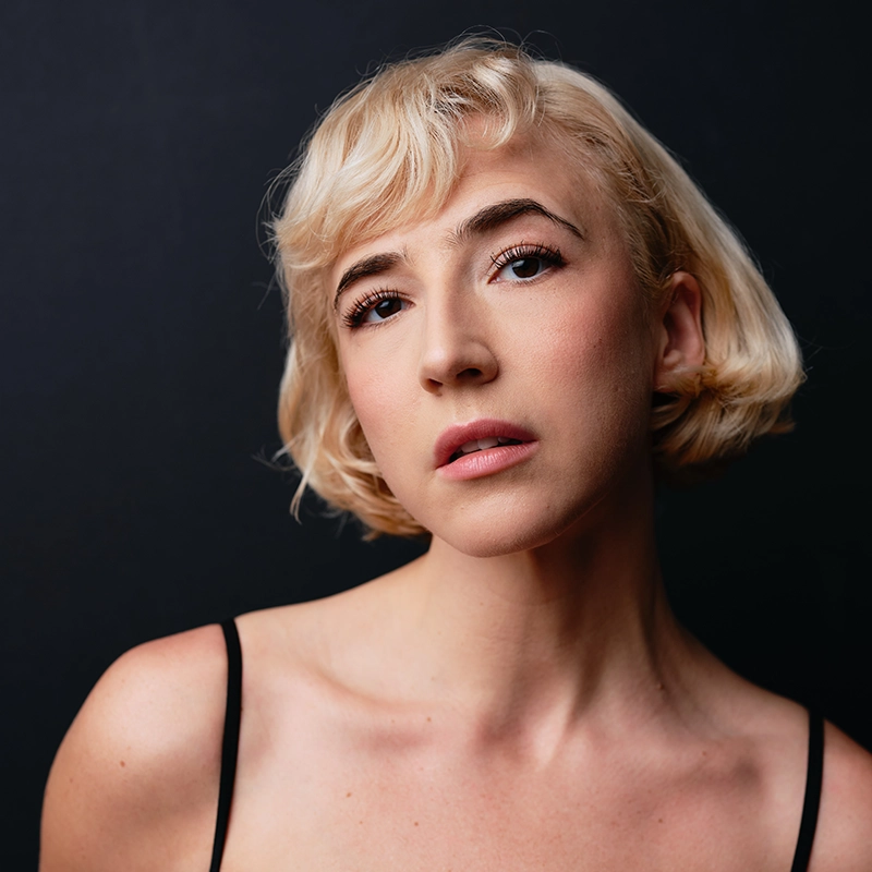 A young person with short, wavy blonde hair and fair skin gazes softly at the camera against a dark background. Reminiscent of Bailey McCall, they wear a black spaghetti strap top, their expression calm and contemplative.