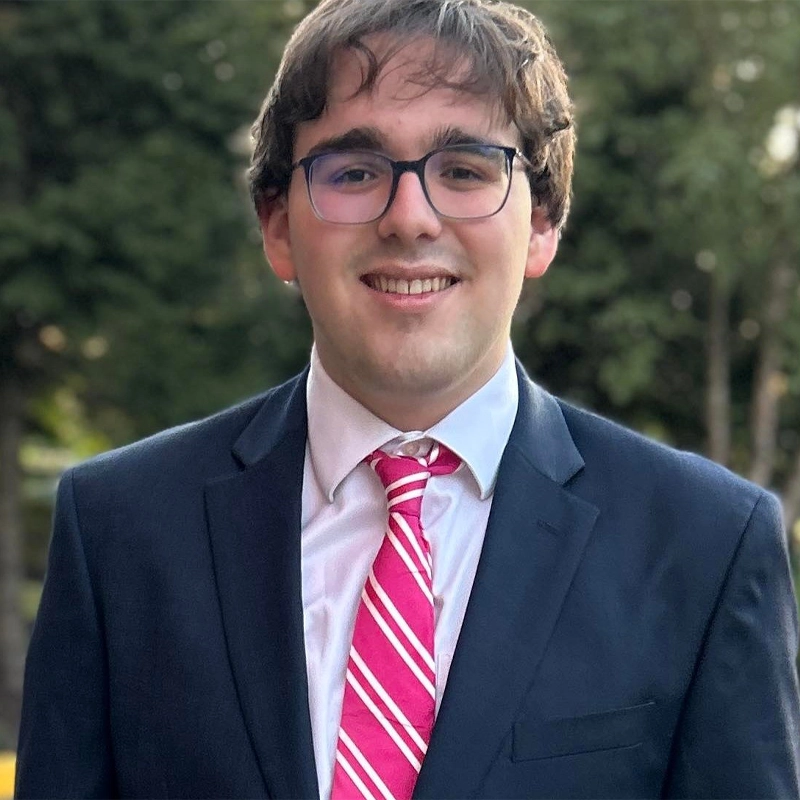A young man with glasses and short brown hair smiles at the camera, wearing a dark suit, a white shirt, and a pink-and-white striped tie. Charles Kirsch stands before trees and greenery in the softly blurred background.